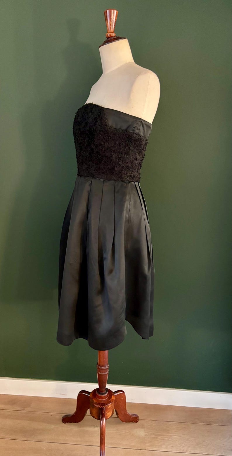 May include: A black strapless dress with a lace bodice and a pleated skirt. The dress is on a wooden mannequin.