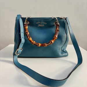 May include: A teal blue leather handbag featuring a textured surface, a curved bamboo handle, and a matching adjustable shoulder strap. The front of the bag displays the gold-toned text MADE IN ITALY BY Gucci, with silver-toned metal hardware accents.