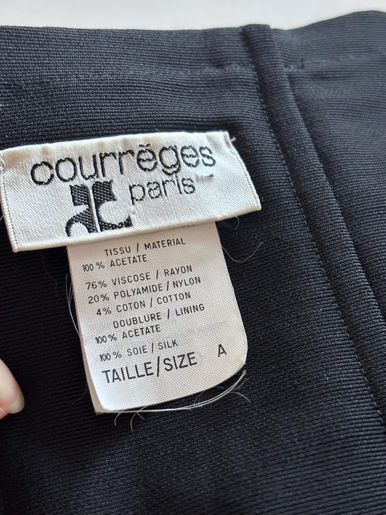 May include: White tag with black text reading "courr&egrave;ges paris" and fabric content information. The tag is attached to a black fabric garment. The fabric content information includes: "TISSU / MATERIAL 100% ACETATE 76% VISCOSE / RAYON 20% POLYAMIDE / NYLON 4% COTON / COTTON DOUBLURE / LINING 100% ACETATE 100% SOIE / SILK TAILLE / SIZE A"