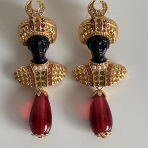 May include: A pair of gold-toned earrings featuring black bust figures with ornate headdresses and jeweled collars. Each earring has a large, teardrop-shaped, red glass pendant. The earrings are set against a white background.