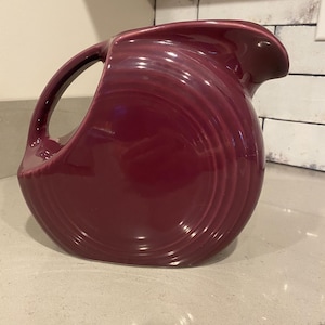 May include: A deep purple ceramic pitcher with a rounded shape and a wide spout. The pitcher has a smooth, glossy finish and a unique, rippled design.