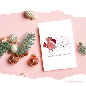 May include: A white greeting card with a cartoon heart wearing a Santa hat and holding an ornament. The card features an EKG line and the text "MERRY QRST-MAS AND A P NEW YEAR!". Christmas ornaments and pine branches are arranged around the card on a pink surface.