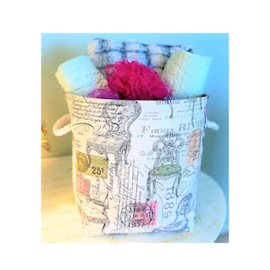 May include: A decorative fabric basket with a vintage Parisian design, filled with rolled towels and a pink loofah. The basket features a cream background with black illustrations of chairs and script. The basket has white rope handles.