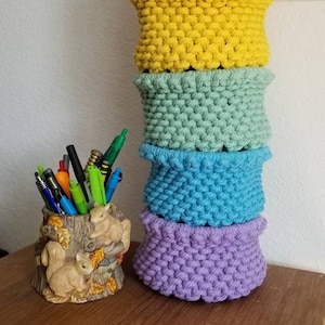 May include: A stack of five colorful crocheted baskets in pink, yellow, green, blue, and purple. The baskets are stacked on a wooden surface.