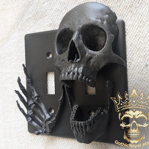 May include: Black light switch cover with a 3D printed skull and skeleton hand design. The skull has an open mouth and the skeleton hand is raised in a gesture of greeting.
