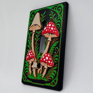 May include: A decorative light switch cover with a relief design of mushrooms. The mushrooms have red caps with white spots and are set against a black background with green foliage. The switch is a standard toggle style.