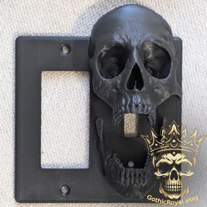 May include: Black skull light switch cover with an open mouth design. The skull is mounted on a black switch plate. A gold skull with a crown is in the lower right corner. The Etsy shop name "GothicRoyal.etsy" is visible.