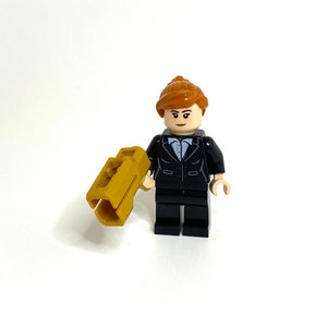 May include: A Lego minifigure of a person in a black suit with a light blue shirt and a gold-colored accessory. The figure has brown hair and is standing on a white surface.