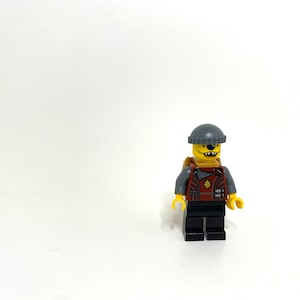 May include: A Lego minifigure with a yellow head, gray beanie, and a red and gray vest. The minifigure has a toothy grin and black pants. The figure is standing on a white surface.