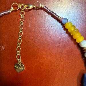 May include: Close-up of a beaded necklace with a gold-colored chain and clasp. The necklace features yellow, white, and blue beads, with a heart-shaped charm that reads "NAKAMOL". The necklace is set against a reddish-brown background.
