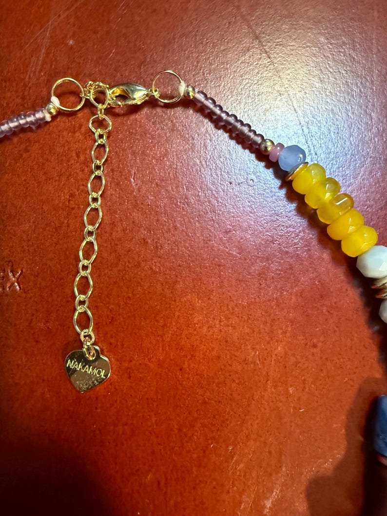 May include: Close-up of a beaded necklace with a gold-colored chain and clasp. The necklace features yellow, white, and blue beads, with a heart-shaped charm that reads "NAKAMOL". The necklace is set against a reddish-brown background.