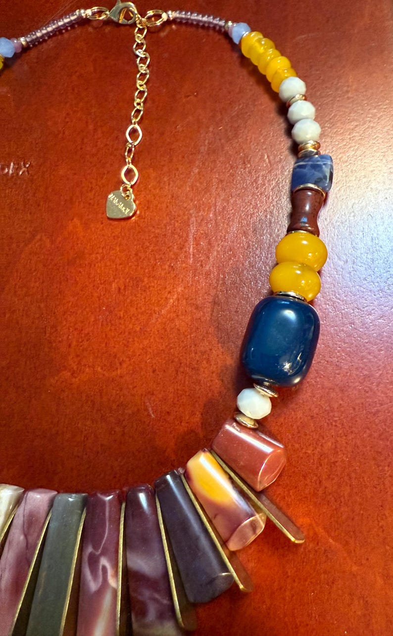 May include: A colorful necklace with a variety of beads in shades of yellow, blue, red, and purple. The necklace has a gold chain and a heart-shaped charm. The beads are various shapes and sizes.