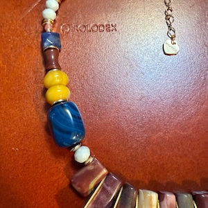 May include: A colorful necklace with a variety of beads in yellow, blue, brown, and white. The necklace has a gold chain and clasp. The text "ROLODEX" is visible on the background.
