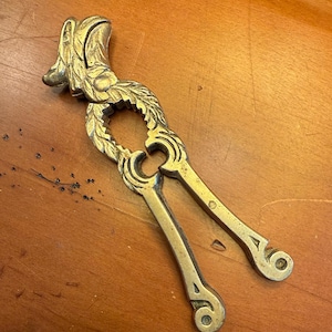 May include: A vintage brass nutcracker with a detailed design. The nutcracker features a decorative head and a leaf-like pattern around the cracking area. The handles have a scrolled design at the ends.