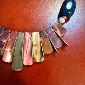 May include: A colorful necklace featuring rectangular, multi-colored stone pendants with gold-colored metal accents. The necklace includes a mix of red, pink, green, and brown stones, along with blue and yellow beads.