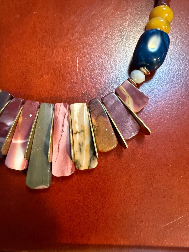 May include: A colorful necklace featuring rectangular, multi-colored stone pendants with gold-colored metal accents. The necklace includes a mix of red, pink, green, and brown stones, along with blue and yellow beads.