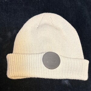 May include: A beige waffle-knit beanie with a folded cuff. Centered on the cuff is a circular grey patch featuring the embossed text Nike Air and the iconic swoosh logo. The hat has a textured, grid-like pattern throughout its construction.