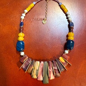 May include: A colorful beaded necklace with a variety of stone and glass beads. The necklace features a mix of yellow, blue, white, and brown beads, with a central section of rectangular, multi-colored stones. The necklace has a gold-colored chain.