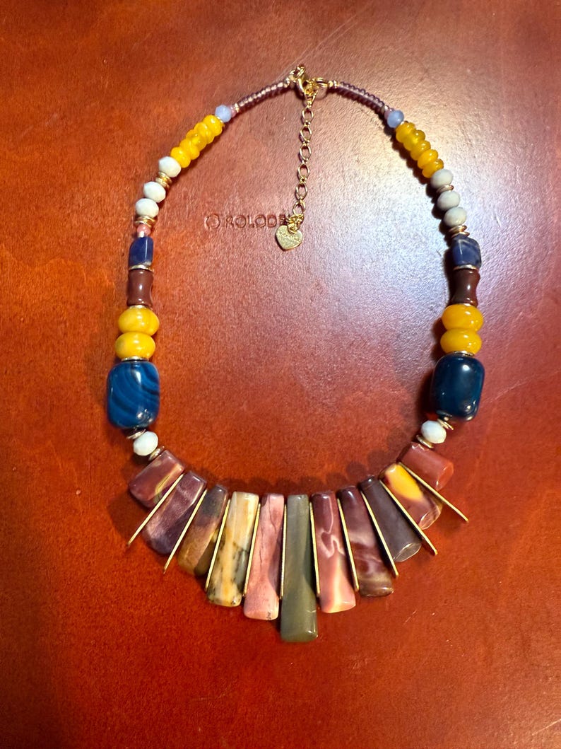May include: A colorful beaded necklace with a variety of stone and glass beads. The necklace features a mix of yellow, blue, white, and brown beads, with a central section of rectangular, multi-colored stones. The necklace has a gold-colored chain.