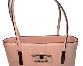 May include: A blush pink leather tote bag with black handles and a gold bow with the text "katespade" on it.