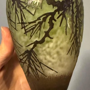 May include: A green and brown glass vase with a pine branch design. The vase has a textured surface and a dark brown base. The pine branches are dark brown and are etched into the glass.
