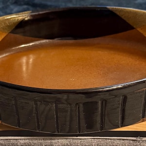 May include: A round ceramic baking dish with a brown interior and a black and gold rim. The exterior has a textured, ribbed design. The dish sits on a wooden surface.