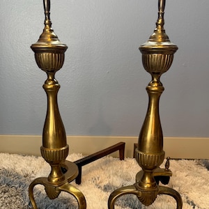 May include: Two antique brass fireplace andirons with ornate detailing. Each andiron features a tall, urn-shaped top, a fluted midsection, and curved legs. A black metal fire grate is visible between the andirons. The andirons are displayed on a patterned rug.