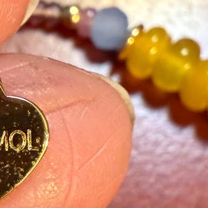 May include: Close-up of a gold heart-shaped charm with the word "NAKAMOL" engraved on it. The charm is attached to a beaded bracelet with yellow and light blue beads. The bracelet is held by a person.