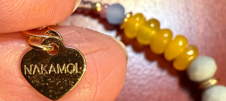 May include: Close-up of a gold heart-shaped charm with the word "NAKAMOL" engraved on it. The charm is attached to a beaded bracelet with yellow and light blue beads. The bracelet is held by a person.