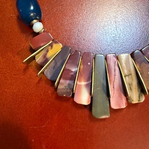 May include: A colorful necklace featuring a combination of large, rectangular, and oval-shaped beads. The necklace has a mix of colors, including red, yellow, blue, and green. The rectangular beads are accented with gold-colored metal.