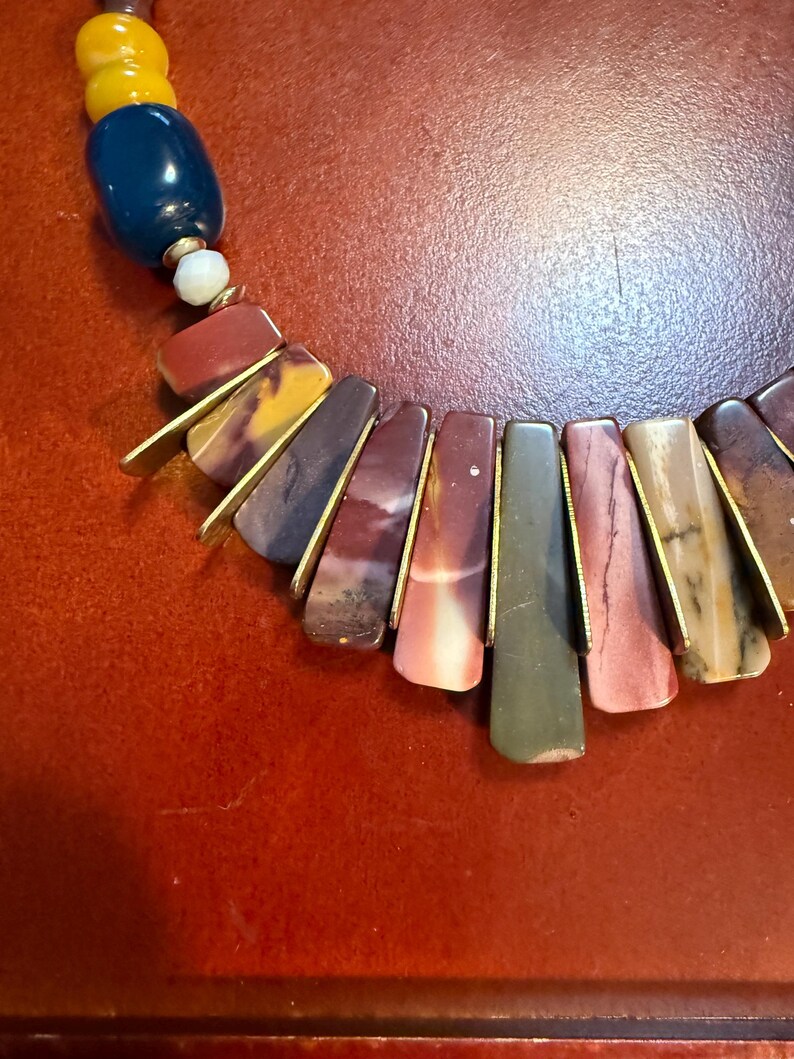 May include: A colorful necklace featuring a combination of large, rectangular, and oval-shaped beads. The necklace has a mix of colors, including red, yellow, blue, and green. The rectangular beads are accented with gold-colored metal.