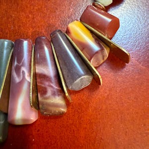 May include: A close-up of a necklace featuring a variety of colorful beads. The necklace includes elongated, teardrop-shaped beads in shades of pink, brown, and gray, accented with gold-colored metal. Other beads are a mix of red, yellow, and white.