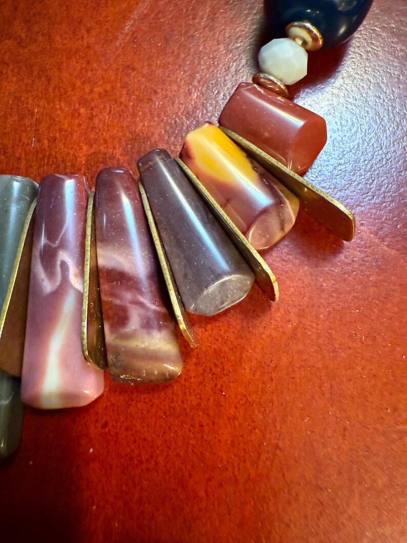 May include: A close-up of a necklace featuring a variety of colorful beads. The necklace includes elongated, teardrop-shaped beads in shades of pink, brown, and gray, accented with gold-colored metal. Other beads are a mix of red, yellow, and white.