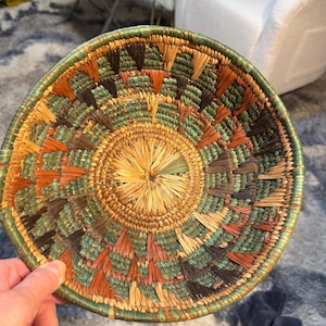 May include: A round, woven basket with a central starburst design. The basket features a green, orange, brown, and natural color palette. The rim is a darker green, and the basket appears to be handmade.
