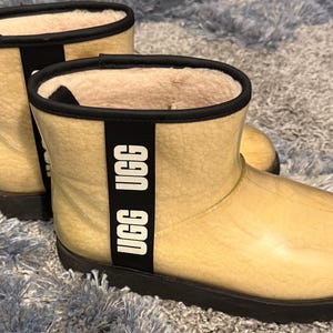 May include: A pair of beige ankle boots with a black sole and trim. The boots have a clear, waterproof exterior and a soft, light-colored interior. A black band with the white text "UGG" is on the side of each boot.