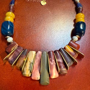 May include: A handcrafted necklace featuring a variety of colorful beads and stones. The necklace includes large, dark blue and yellow beads, along with smaller, multi-colored beads. The focal point is a series of rectangular stones in shades of red, brown, and green, accented with gold-colored metal.