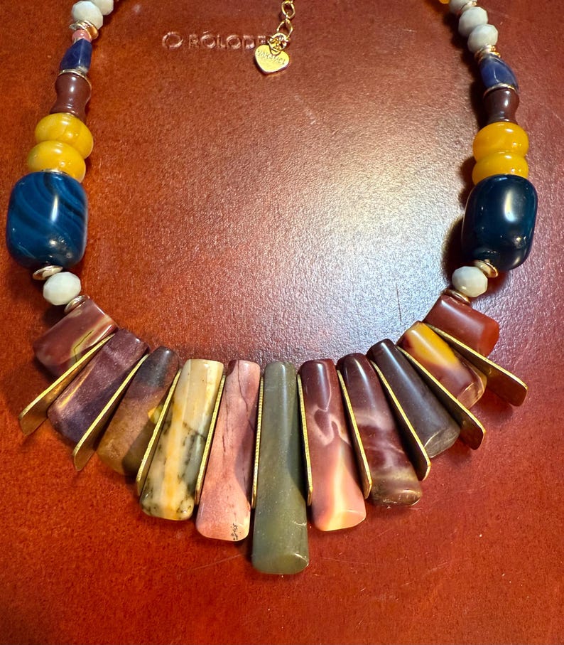 May include: A handcrafted necklace featuring a variety of colorful beads and stones. The necklace includes large, dark blue and yellow beads, along with smaller, multi-colored beads. The focal point is a series of rectangular stones in shades of red, brown, and green, accented with gold-colored metal.