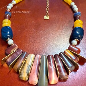 May include: A colorful necklace featuring a variety of beads in shades of yellow, blue, brown, and pink. The necklace has a gold-tone chain and pendant. The necklace is approximately 18 inches long.