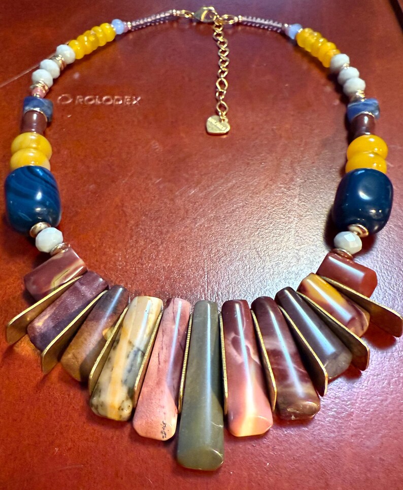 May include: A colorful necklace featuring a variety of beads in shades of yellow, blue, brown, and pink. The necklace has a gold-tone chain and pendant. The necklace is approximately 18 inches long.