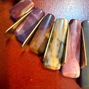 May include: A close-up of a necklace featuring a variety of colorful, rectangular stone beads. The beads are held in place by gold-colored metal accents. The necklace also includes round beads in white, gold, and dark blue.