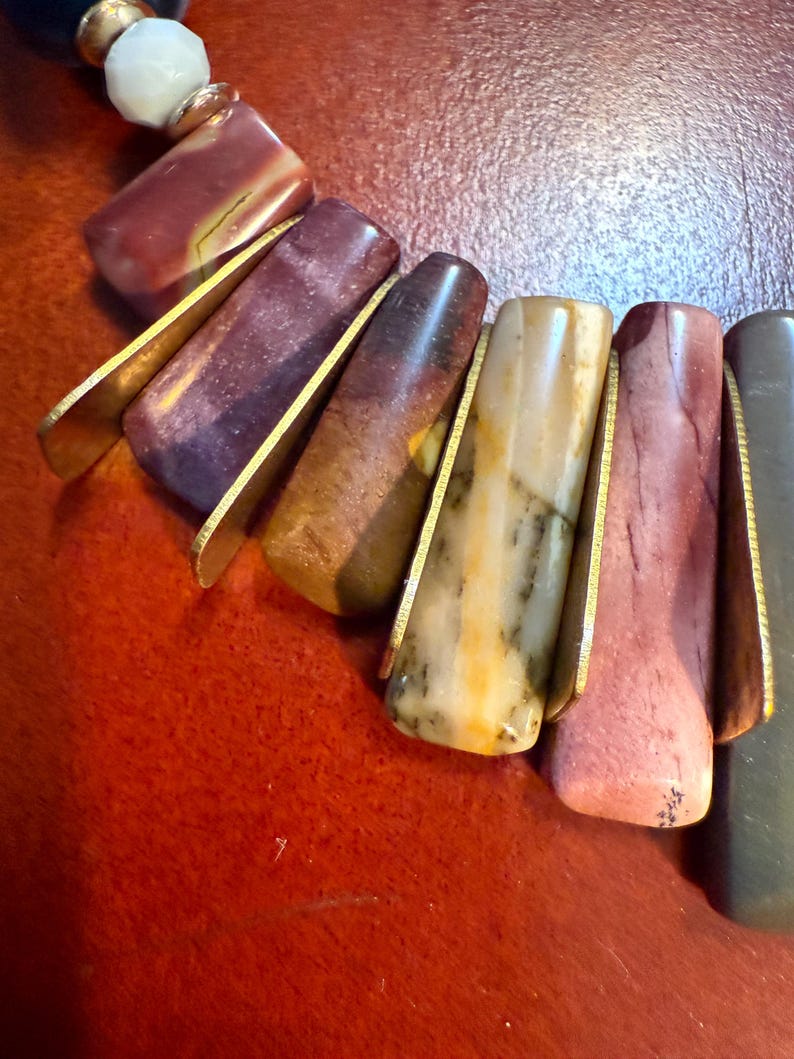 May include: A close-up of a necklace featuring a variety of colorful, rectangular stone beads. The beads are held in place by gold-colored metal accents. The necklace also includes round beads in white, gold, and dark blue.