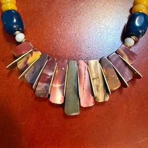 May include: A colorful necklace featuring a variety of stone beads in shades of blue, yellow, and burgundy. The focal point is a fan-shaped arrangement of rectangular stones in earthy tones, accented with gold-colored metal. A small gold heart charm is visible.
