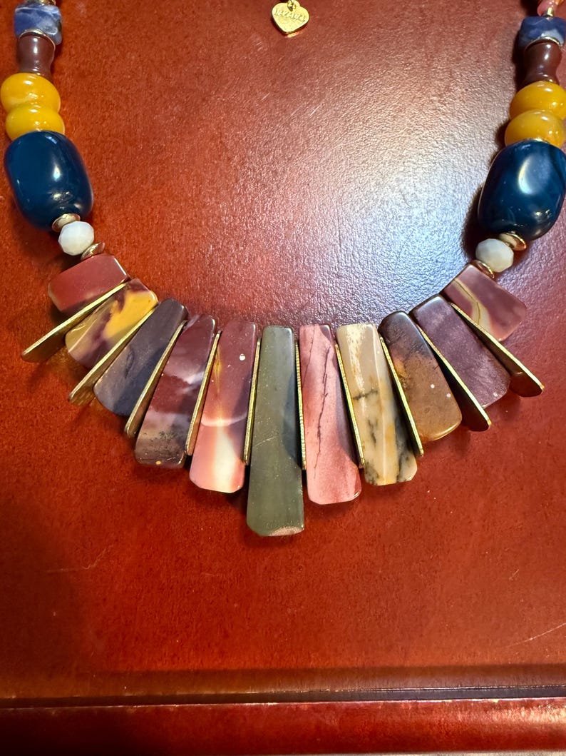 May include: A colorful necklace featuring a variety of stone beads in shades of blue, yellow, and burgundy. The focal point is a fan-shaped arrangement of rectangular stones in earthy tones, accented with gold-colored metal. A small gold heart charm is visible.