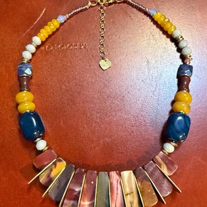 May include: A colorful necklace featuring a variety of beads in shades of yellow, blue, brown, and red. The necklace has a gold-colored chain and clasp. The focal point is a series of rectangular, multi-colored stone pendants.