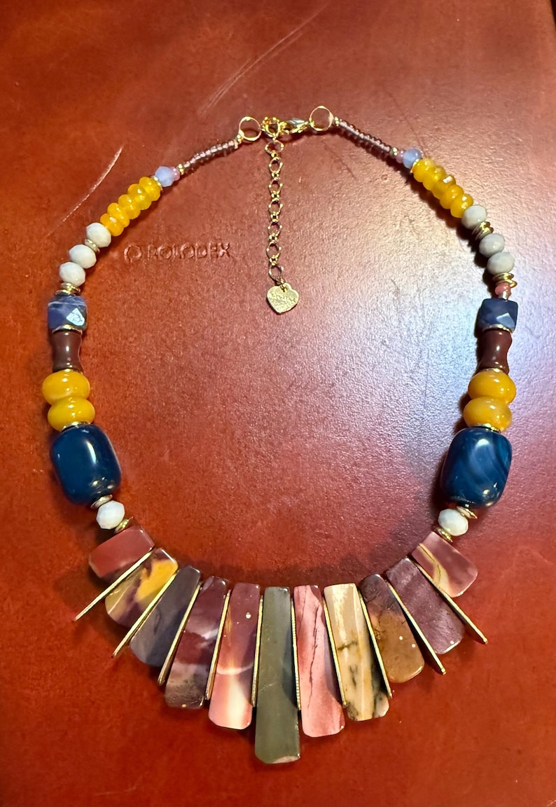 May include: A colorful necklace featuring a variety of beads in shades of yellow, blue, brown, and red. The necklace has a gold-colored chain and clasp. The focal point is a series of rectangular, multi-colored stone pendants.