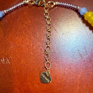 May include: Close-up of a gold-toned necklace clasp and extender chain. The necklace features purple, blue, and yellow beads. A heart-shaped charm with the brand name "NAKAMOL" is attached to the end of the extender chain.