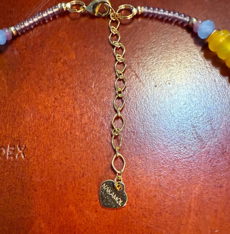 May include: Close-up of a gold-toned necklace clasp and extender chain. The necklace features purple, blue, and yellow beads. A heart-shaped charm with the brand name "NAKAMOL" is attached to the end of the extender chain.