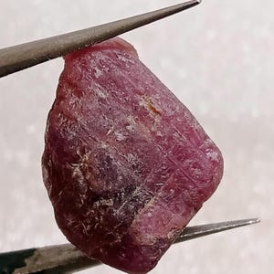Könnte beinhalten: Ein roher, unpolierter Rubin-Edelstein mit einem tiefen Magenta- und Dunkelrot-Farbton. Der Stein weist eine natürliche, unregelmäßige Form mit sichtbaren kristallinen Texturen, rauen Oberflächen und inneren Einschlüssen auf.
