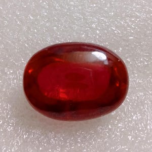 Könnte beinhalten: Ein leuchtend roter, oval geformter Edelstein mit einer glatten, polierten Cabochon-Oberfläche. Der Stein zeigt einen tiefen, durchscheinenden karmesinroten Farbton mit subtilen internen Lichtreflexionen und einem glänzenden Finish.