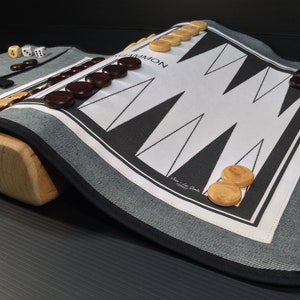 Pode incluir: Um conjunto de backgammon com uma superfície de jogo a preto e branco, dados e peças de jogo de madeira. O tabuleiro está enrolado e é mantido no lugar por um suporte de madeira. As peças são castanhas e de madeira natural.